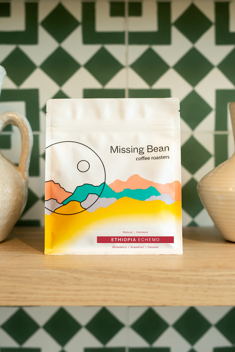 A bag of Missing Bean's Ethiopia Echemo on a wooden shelf between two vases against a green patterned tiled wall.