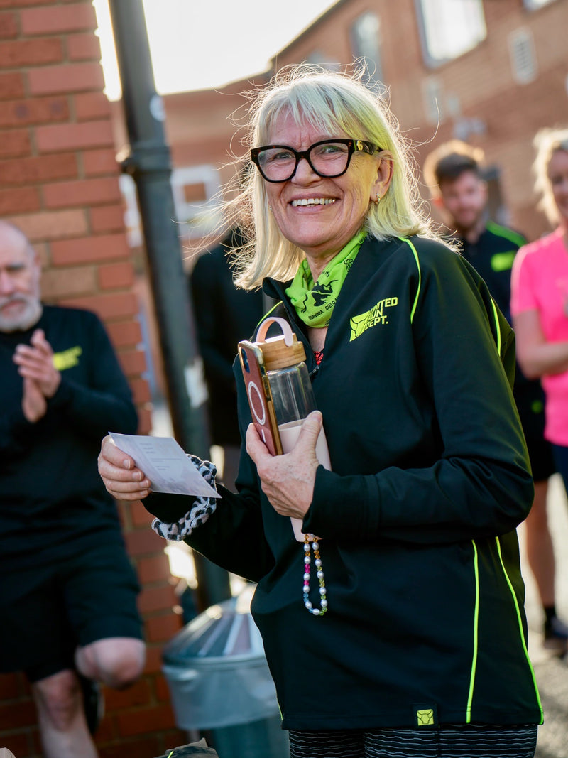 Woman in an Oxford United Run Department black jacket holding a water bottle and phone, standing outdoors.