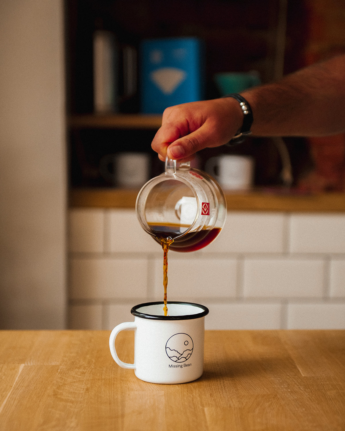 A hand pouring coffee from a glass coffee pot into a white Missing Bean branded mug on a wooden table at the Missing Bean coffee shop in Banbury