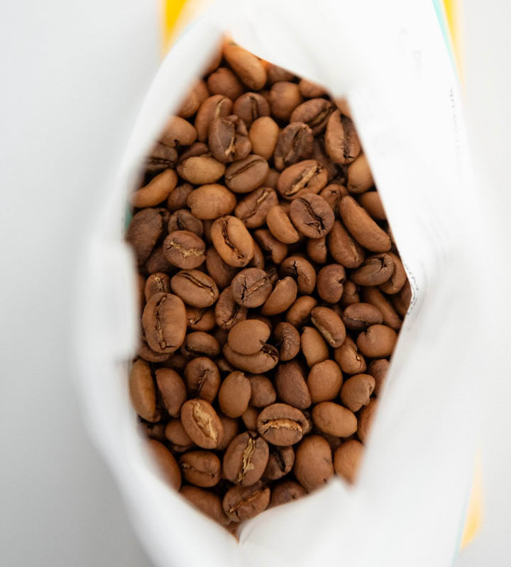  An overhead view captures the inside of a bag of Missing Bean Coffee beans, with the beans sharply in focus. The rest of the bag, showcasing some of the vibrant colors of Missing Bean's branding, is blurred in the background. The bag is placed on a white table.