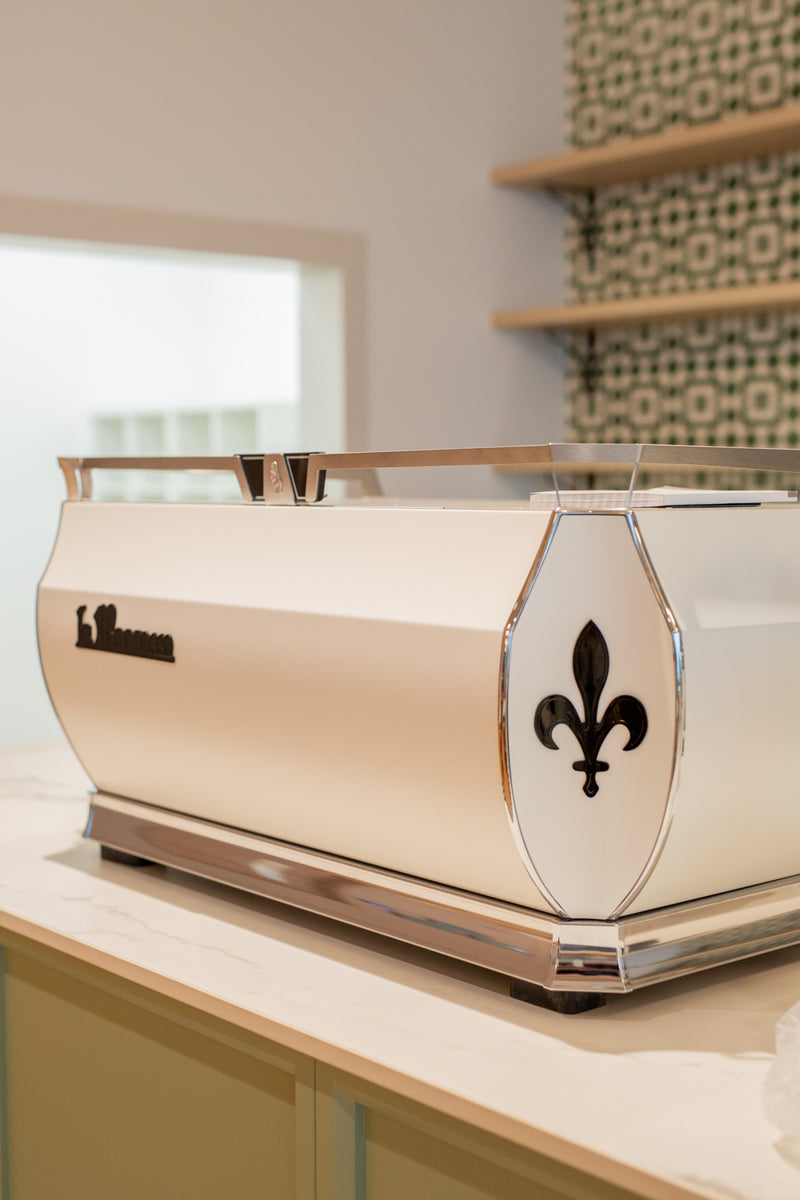 A white La Marzocco coffee machine with a black fleur de lys on a wooden surface.