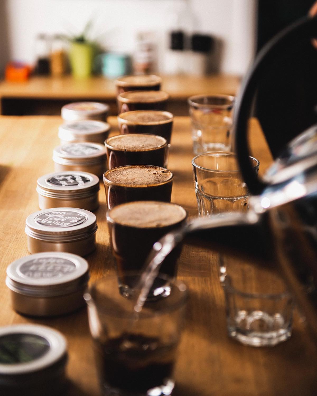 A cup of coffee beans being filled up with water, in line on a wooden table with other full cups, ready for a coffee cupping session.