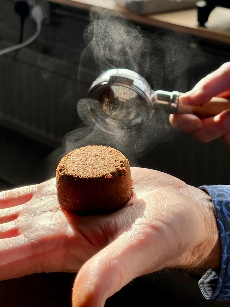 A spent espresso puck resting on an open palm beside a steaming portafilter, backlit by sunlight.