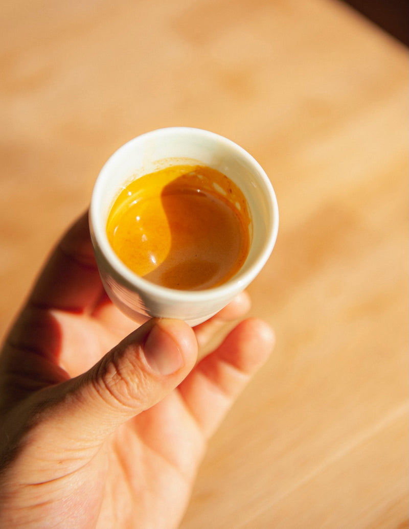 A hand holding a small ceramic cup of freshly pulled espresso with a rich golden crema, shot from above in warm sunlight.