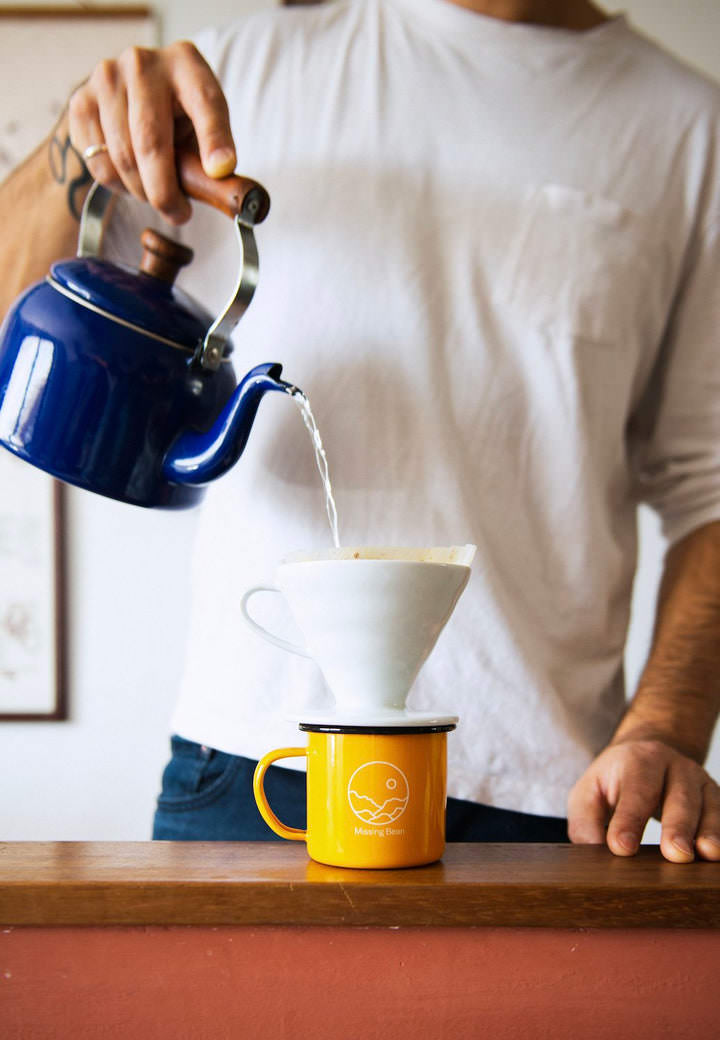 A man in a white t shirt and blue jeans with a wrist tattoo and a silver ring on his pinky pours water out of a navy blue kettle into a white Hario V 60 with a filter which is sitting ontop of a yellow Missing Bean enamle mug, which as a black lip and has a white Missing Bean logo positioned to the front. It is clear from the filter that there are Missing Bean coffee grounds in the V60. 