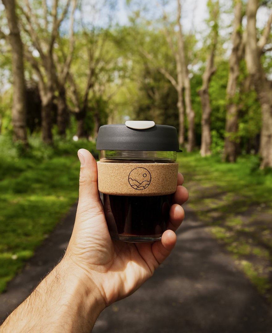 A man's hand holds a Missing Bean Keep Cup, featuring a black plastic lid and a cork band adorned with the black Missing Bean logo. The cup's transparent glass body reveals the dark coffee inside. Against the backdrop of a woodland path, with lush green grass, trees, and a blue sky, the cup is held, the surroundings softly blurred.