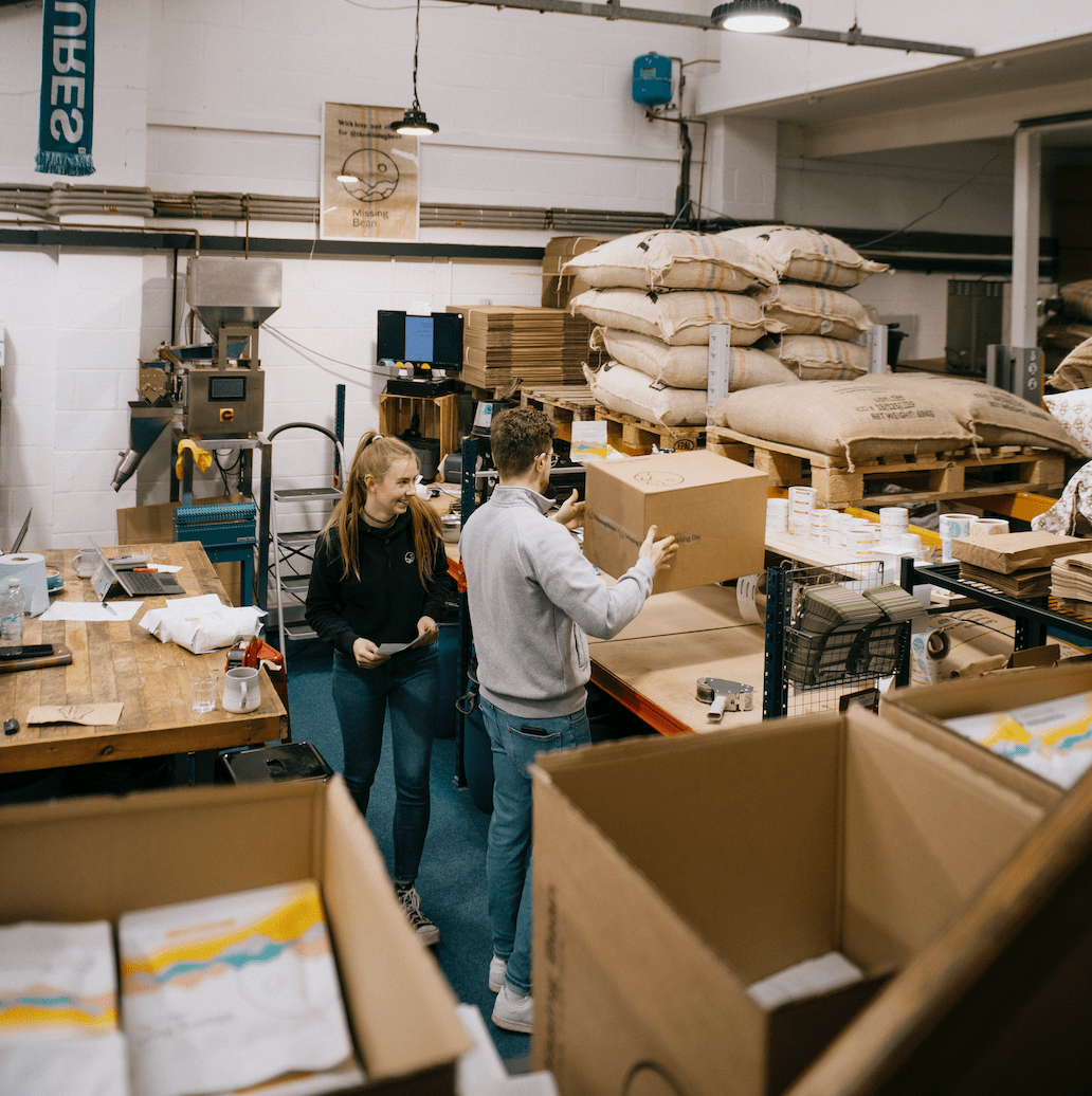 Two people in a warehouse filled with coffee roasting equipment and sacks of roasted coffee beans, work on packaging deliveries of wholesale coffee beans in Oxford.