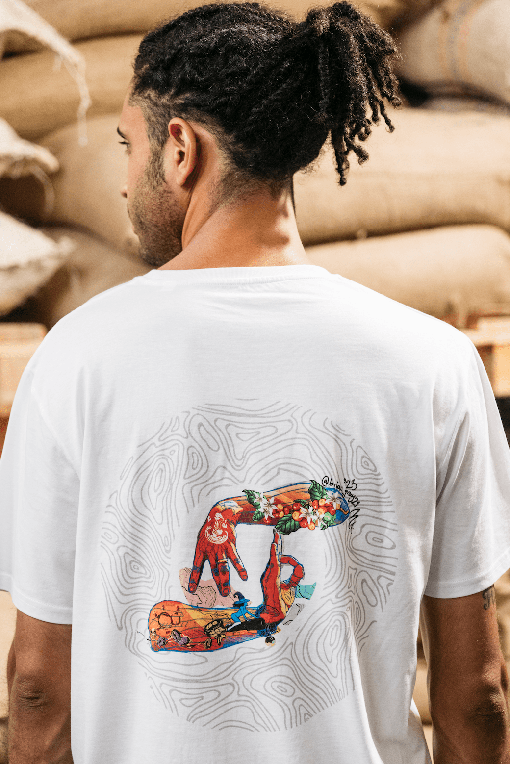 A person in front of stacked burlap sacks of coffee beans. They are wearing a white Missing bean X Brian Gray T-Shirt featuring a colorful graphic design on the back, depicting two hands spelling out MB which are detailed with a range of clever, original, sketches to signify the journey of a coffee bean.