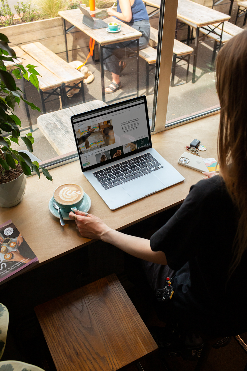 A person with long hair wearing a black t shirt holds a blue coffee cup filled with latte art, looking at Missing Bean's online shop on a laptop. They are in the window of Missing Bean's East Oxford Roastery.