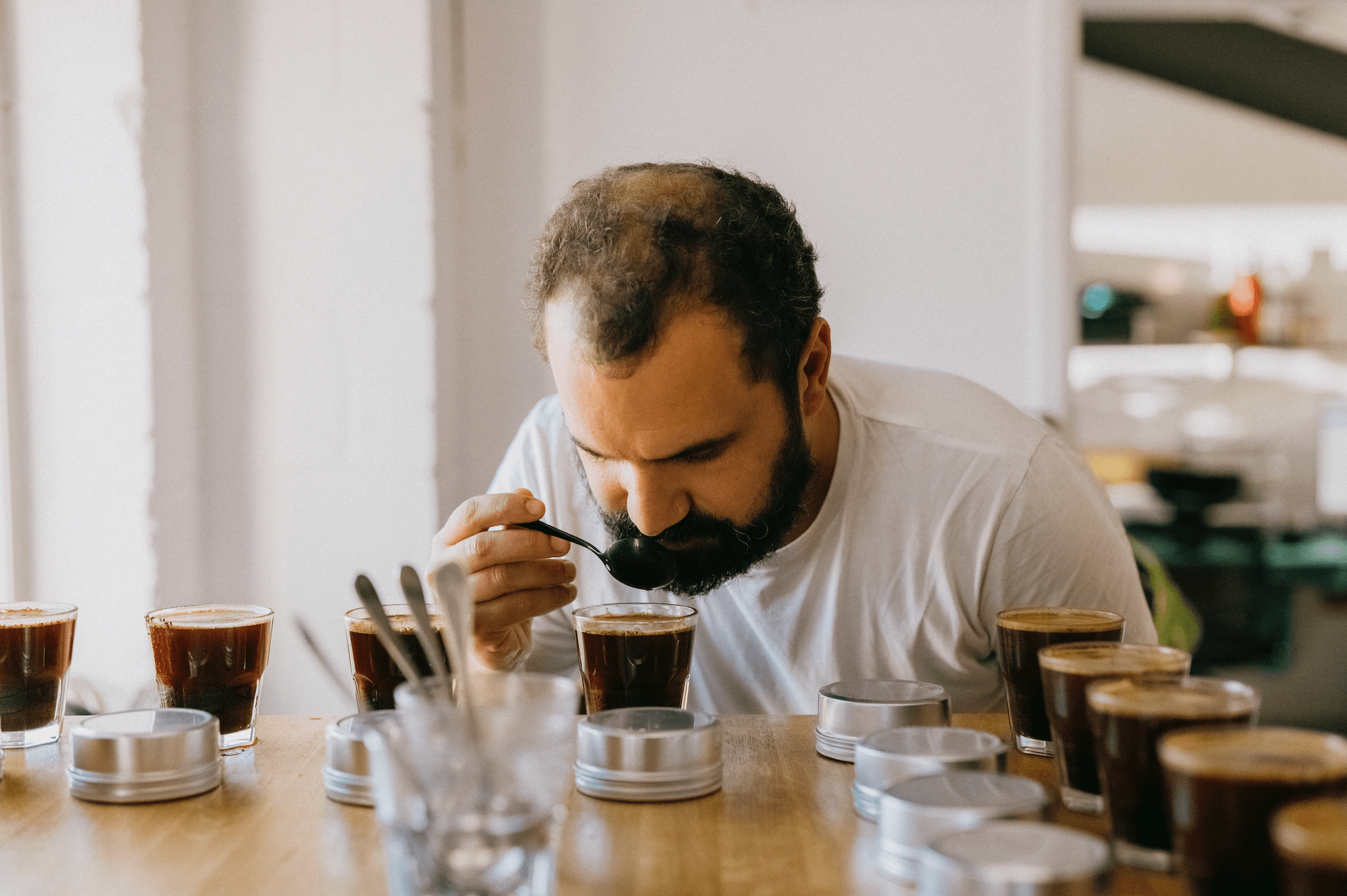 A man performs a coffee cupping session, tasting coffee samples with a spoon at a wooden table.