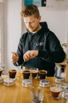 A person wearing a black hoodie with a Missing bean coffee roastery logo concentrates on spooning coffee grounds into a clear glass container. Several other cups and silver lids are on the wooden table in front of them, as part of the Missing Bean coffee tasting session.