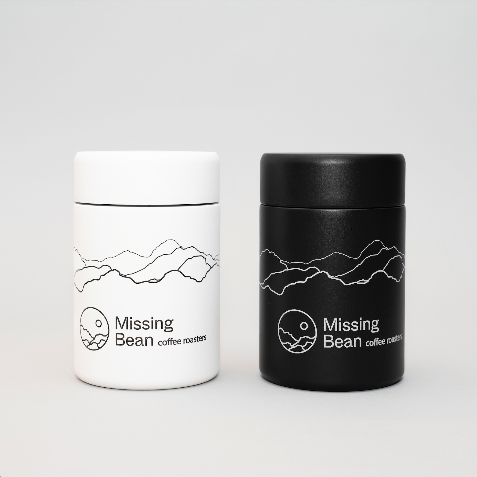 A white and a black coffee container from Missing Bean coffee roasters. Both feature minimalist line art of a mountainous landscape with a rising or setting sun on their labels and the Missing Bean logo. The white container has black artwork, and the black container has white artwork.