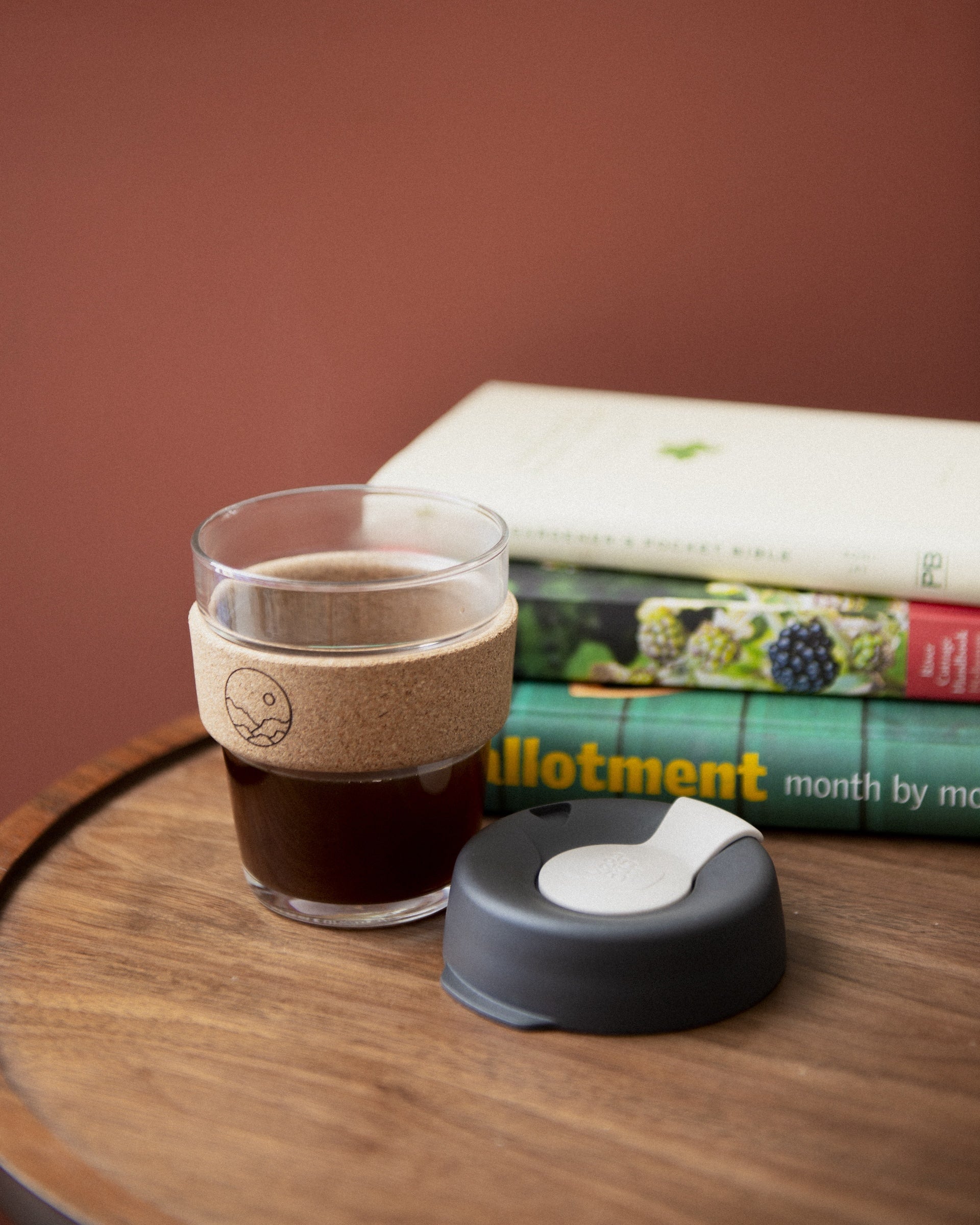 A glass takeaway coffee cup with a black lid and a cork heat-proof band placed on a wooden table with books in the background.